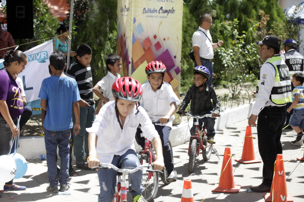 Kinder fahren Fahrräder auf einer Straße mit Verkehrskegeln, einige tragen Helme, andere stehen daneben, mit einer Fahne, Bäumen und Gebäuden im Hintergrund.