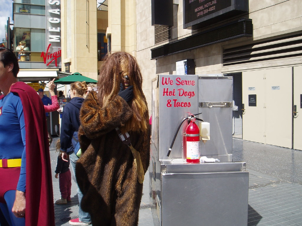 Eine Menschenmenge, einige in Kostümen, versammelte sich vor einem Gebäude mit sichtbarer Beschilderung und Türen, mit einer Kiste und einem Feuerlöscher in der Nähe.