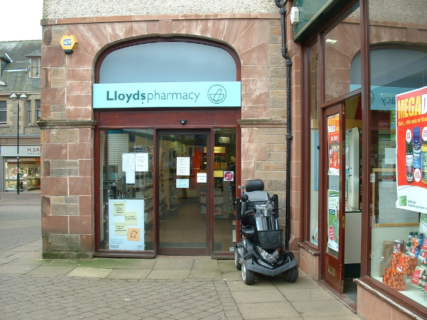 Eine Apothekenfiliale mit einem vor ihr geparkten Fahrzeug und einem Gebäude, das links sichtbar ist.