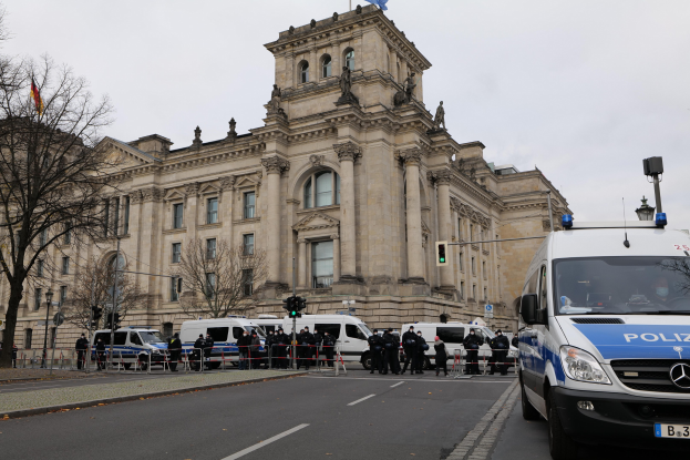 Eine Gruppe von Polizisten vor dem Reichstaggebäude in Berlin, Deutschland, mit Fahrzeugen, einem Zaun, Verkehrszeichen, Laternenpfählen, Bäumen und Flaggen im Hintergrund.