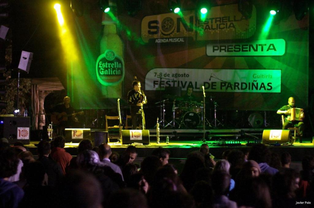 Ein Publikum schaut einer Performance auf der Bühne mit Musikern, die Instrumente spielen, Lautsprechern, Mikrofonen, Werbetafeln und fokussiertem Licht zu, wobei eine Flasche und andere Gegenstände im Hintergrund sichtbar sind.