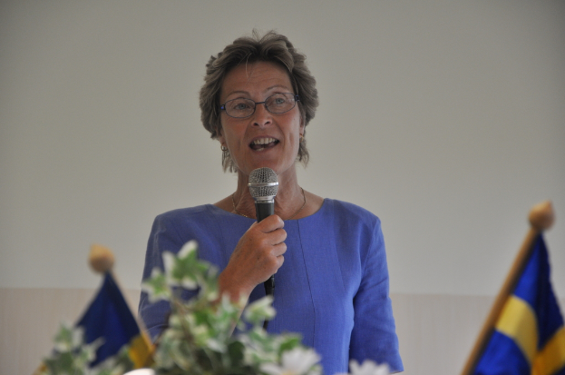 Eine Frau steht an einem Rednerpult mit zwei Flaggen und spricht in ein Mikrofon.