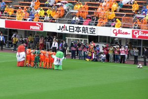 Ein Fußballspiel in einem Stadion mit sechs Spielern, drei Fußballen, vielen Zuschauern in Regenmänteln, die Regenschirme halten, und mehreren Kameramännern.