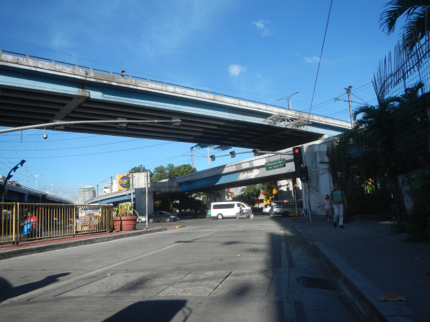 Eine Stadtstraße mit einer Fußgängerbrücke darüber, Fahrzeuge, Fußgänger, Schilder, Laternen, Bäume und Gebäude unter einem bewölkten Himmel.