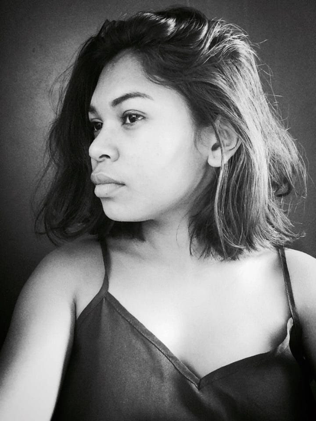 Eine selbstbewusste Frau in einem schwarzen Kleid posiert in einem Schwarz-Weiß-Foto, das sie vor einer Wand mit einem glatten Bob und natürlichem Make-up zeigt.