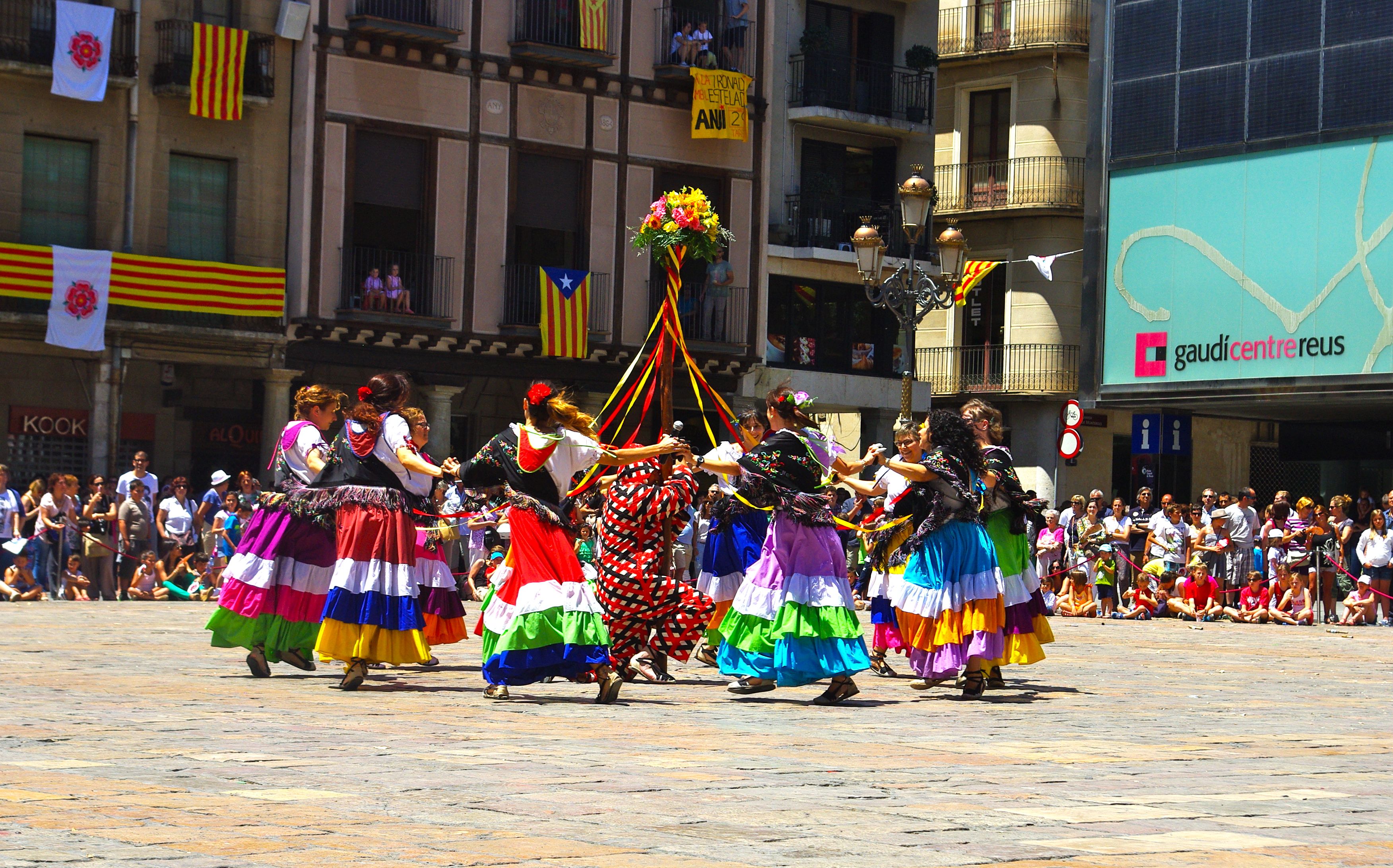 Eine Gruppe von Frauen in bunten Kleidern, die auf der Straße tanzen, umgeben von einer Menge Menschen, mit Gebäuden, Geländern, Balkonen, Fahnen, einem Laternenmast und einer Tafel mit Schrift im Hintergrund.
