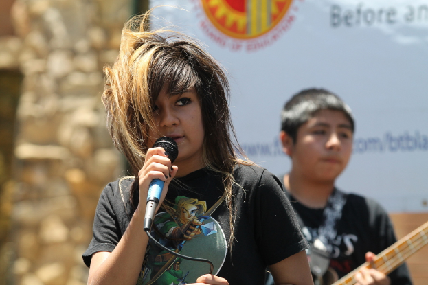 Ein Mädchen hält ein Mikrofon, während ein Junge hinter ihr auf der Bühne eine Gitarre spielt.