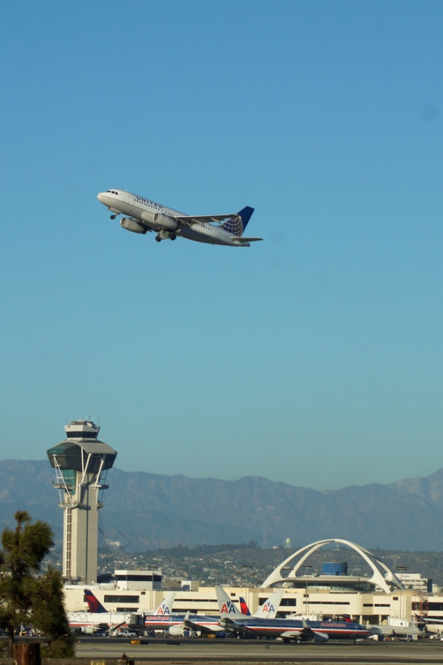 Blick auf einen Flughafen mit einem weißen Kontrollturm im Vordergrund, parkenden Flugzeugen auf dem Boden, entfernten Gebäuden und einem Flugzeug am Himmel.