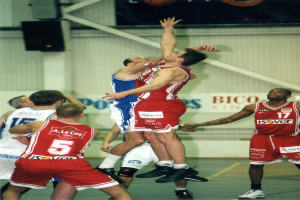 Eine Gruppe von Männern, die Basketball auf einem Court mit einer Wand spielen, auf der Text und Lautsprecher zu sehen sind, und ein Basketball in der Luft an der Spitze des Bildes.
