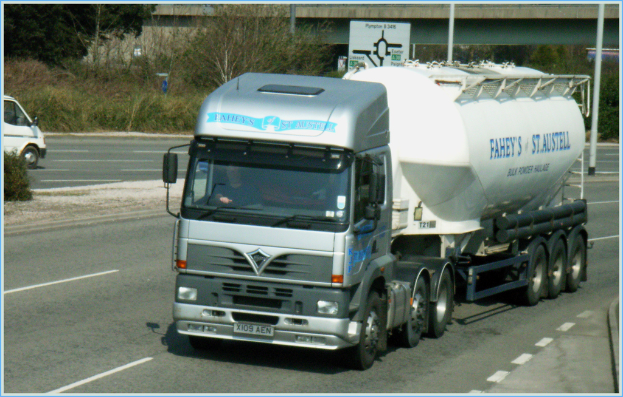 Ein Lkw mit Fahrer fährt auf einer Straße, mit einem Van auf der Gegenfahrbahn, Bäumen und einer Brücke im Hintergrund.