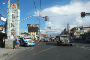 Eine belebte Stadtstraße mit Fahrzeugen, Fußgängern, Gebäuden, Ampeln, Strommasten, Bäumen und einem bewölkten Himmel.