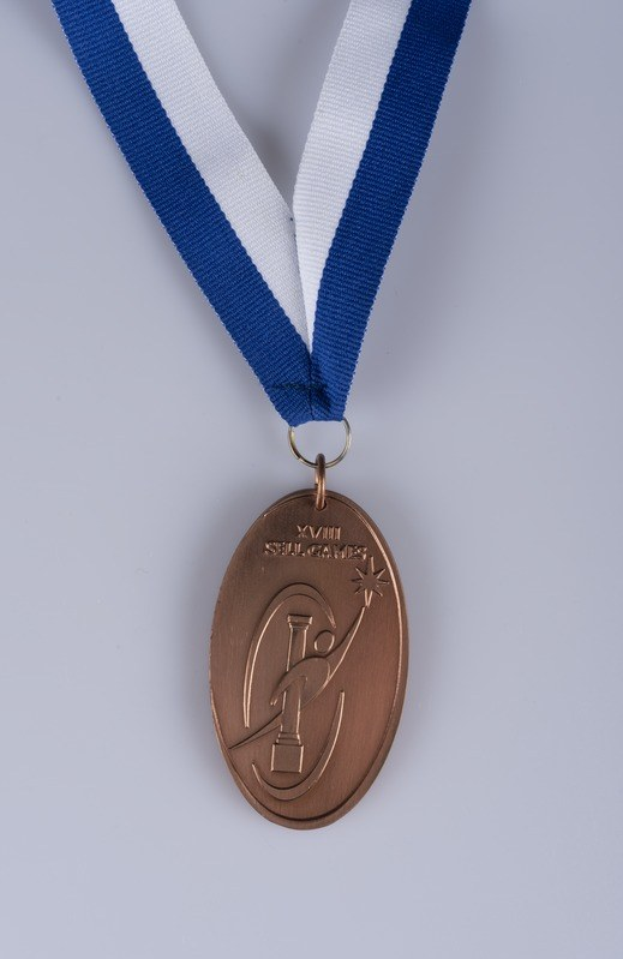 Goldmedaille mit einem blauen und weißen Band auf einem weißen Hintergrund, mit eingravierter Beschriftung, die darauf hinweist, dass es eine Sportsmedaille ist.