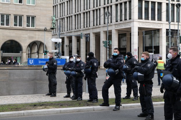 Polizeibeamte in schwarzen Uniformen und Masken vor einem Gebäude mit Glasfenstern und Säulen in Berlin, Deutschland, einige mit Helmen, mit Laternen, Verkehrsampeln, Schildern mit Text, einer Statue und anderen Menschen im Hintergrund.
