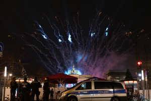 Ein Polizeiwagen parkt vor einer Menschenmenge, mit Fahrrädern, Zelten, Laternenmasten, Schildern, Verkehrszeichen, Bäumen, Gebäuden und einer Statue im Hintergrund, unter einem Himmel, der von Feuerwerk erhellt wird.