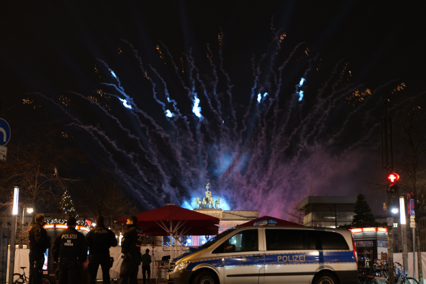 Ein Polizeiwagen parkt vor einer Menschenmenge, mit Fahrrädern, Zelten, Laternenmasten, Schildern, Verkehrszeichen, Bäumen, Gebäuden und einer Statue im Hintergrund, unter einem Himmel, der von Feuerwerk erhellt wird.