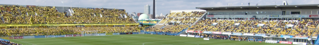 Großes Stadion voller Zuschauer bei einem Fußballspiel auf saftigem Grün, umgeben von Werbetafeln, mit Gebäuden, Pfählen und einem klaren blauen Himmel im Hintergrund.