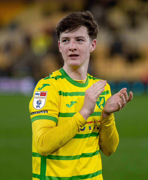 Ein junger Mann in einem gelb-grünen T-Shirt steht auf einem Fußballfeld und klatscht begeistert in die Hände, mit einem unscharfen Hintergrund.