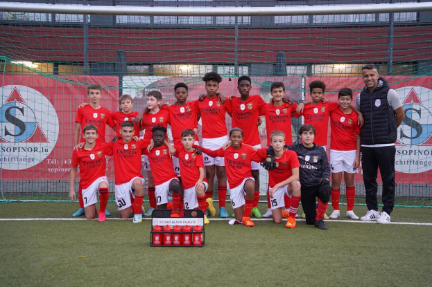 Eine Gruppe junger Männer in roten T-Shirts und schwarzen Shorts steht auf einem Fußballfeld neben einer Kiste roter Gegenstände, im Hintergrund sind Schilder mit der Aufschrift "FC Rot-Schwarz Eglaute - Champions League" zu sehen.