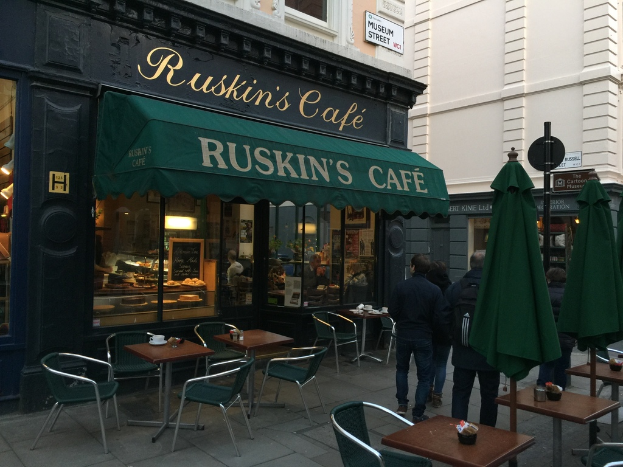 Eine Gruppe von Menschen steht vor einem Café mit einer Tafel, auf der "Ruskin's Café" steht.