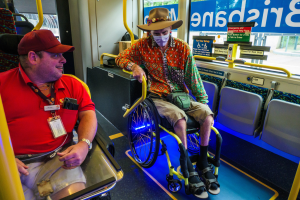 Zwei Männer im Rollstuhl in einem Bus, einer trägt ein rotes T-Shirt und eine Kappe, der andere ein orangefarbenes Hemd und einen braunen Hut, beide tragen Masken und Ausweise, mit gelben Stäben und Glasfenstern, durch die man Bäume, Gebäude und Himmel sieht.