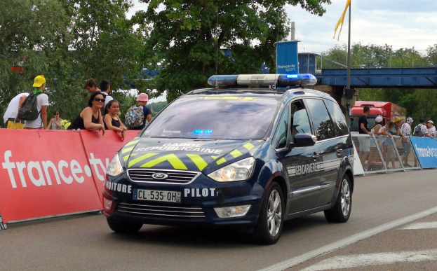Polizeiauto fährt auf einer Straße neben einer Menge, mit einer Fahne links, Geländern mit Fahnen dahinter, Bäumen, einer Brücke, einer Fahne an einem Mast und einem bewölkten Himmel im Hintergrund.