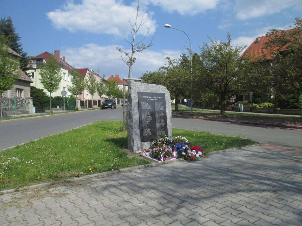 Ein Gedenkstein in einer Stra{\ss}e umgeben von Gras, Pflanzen, B"{a}umen, Laternenmasten und Fahrzeugen mit Blumen darauf, gewidmet den Opfern des Holocaust, mit Geb"{a}uden und einem bew"{o}lktem Himmel im Hintergrund.