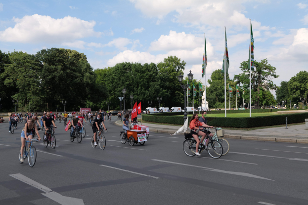 Eine Gruppe von Menschen, die auf Fahrrädern eine von Bäumen gesäumte Straße mit Laternenmasten und Flaggenmasten entlangfahren, während sie an der Pariser Fahrradtour 2016 teilnehmen.