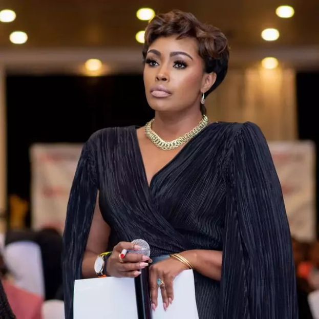 Eine Frau in einem schwarzen Kleid hält ein Mikrofon und Papier und diskutiert mit einem sitzenden Publikum unter Deckenbeleuchtung.