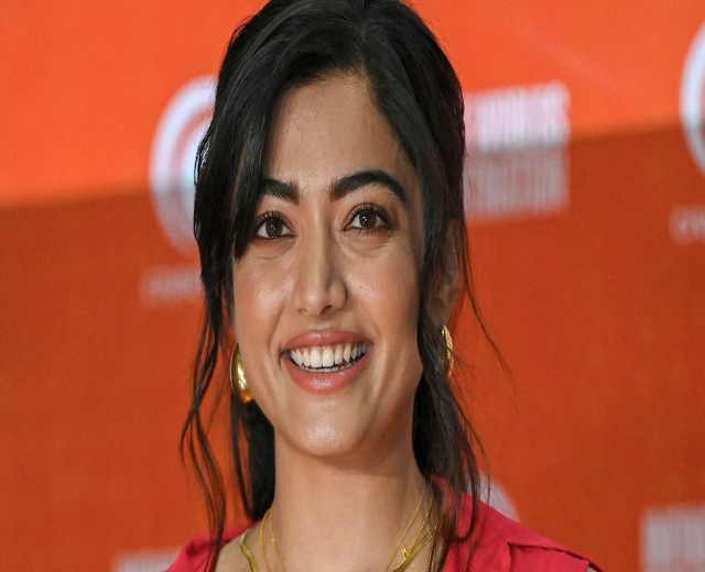 Eine Frau in einem roten Kleid mit einem eleganten Dutt und dezentem Make-up steht strahlend da, trägt schwarze High Heels und hat ein hellgelbes Banner mit der Aufschrift 'Die besten Schönheits Looks von Sonam Kapoor beim Cannes Film Festival' im Hintergrund.
