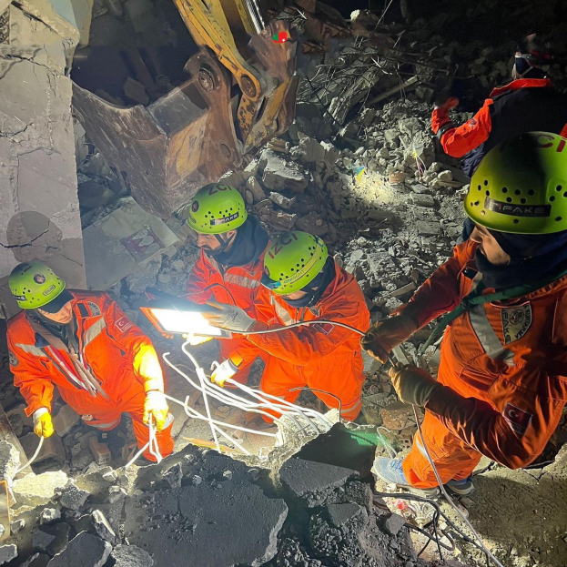 Gruppe von Menschen in orangen Uniformen und grünen Helmen bei der Arbeit an Schutt mit einem Kran im Hintergrund während eines Einsatzes nach dem Einsturz eines Gebäudes.