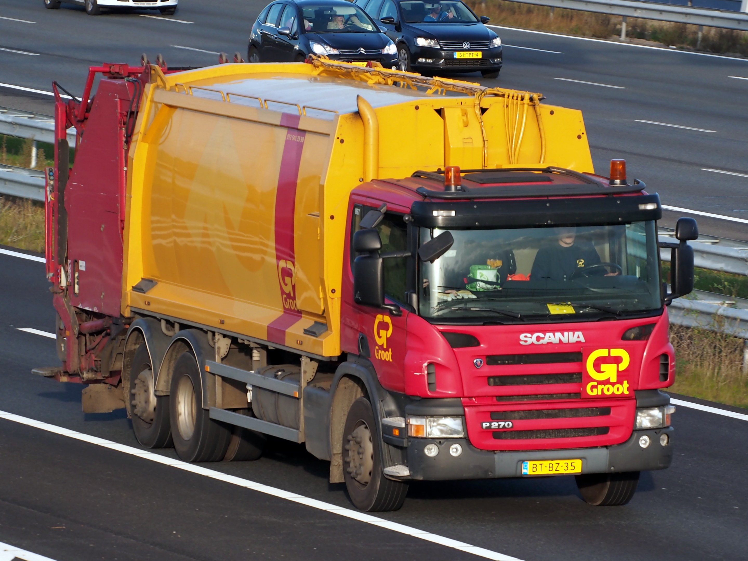 Gelber und roter Müllwagen auf einer Autobahn, umgeben von Autos, mit einer Begränzung und Gras auf beiden Seiten der Straße.