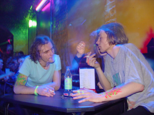 Ein Tisch mit einer Flasche, einer Speisekarte und einem Aschenbecher in der Mitte eines Innenraums, mit zwei Männern und einer Frau, die Zigaretten rauchen.