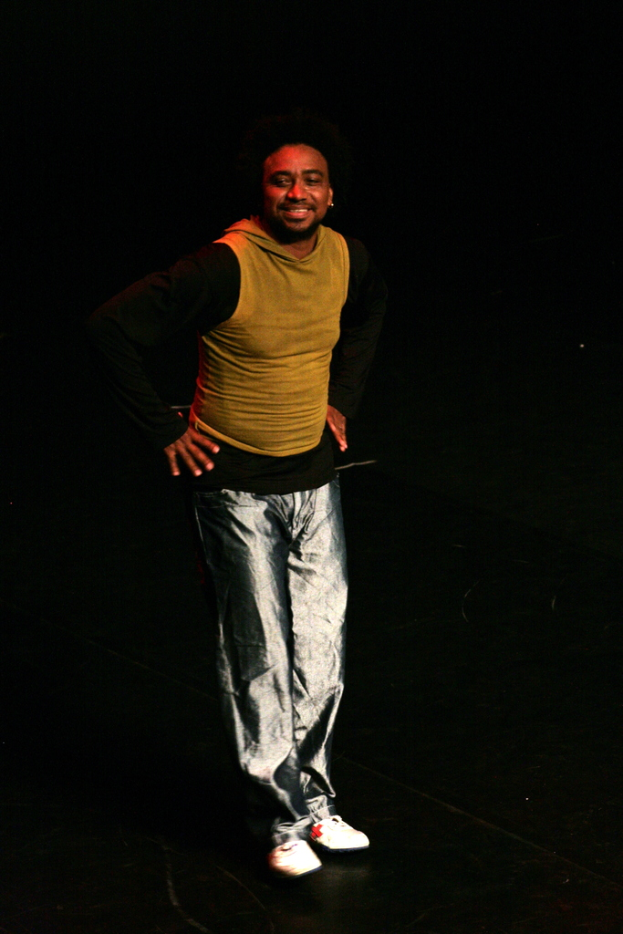 Eine Person in einem T-Shirt lächelt beim Auftritt auf einer Bühne mit dunklem Hintergrund.