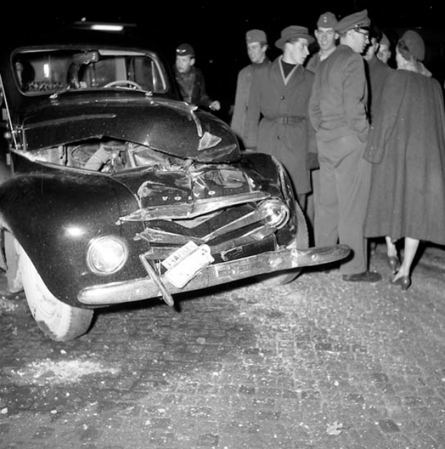 Eine Schwarz-Weiß-Szene eines beschädigten Autos am Straßenrand, mit einer Gruppe von Menschen drumherum, die besorgt aussehen.