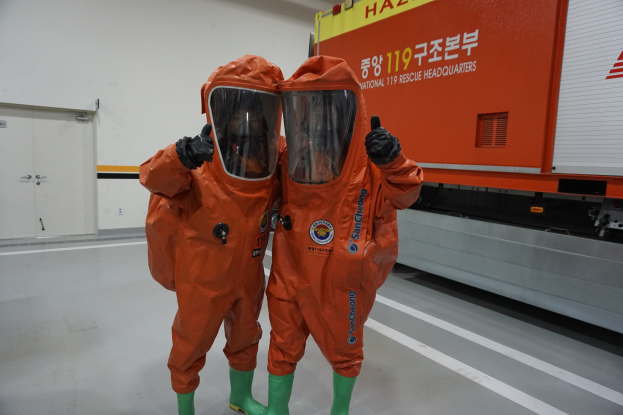 Zwei Personen in orangefarbenen Schutzanzügen stehen nebeneinander vor einem Truck, mit einer Wand und Türen im Hintergrund, was darauf hinweist, dass sie Teil des Nationalen Einsatzteams sind.