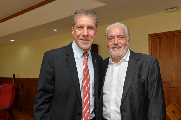 Zwei Personen in Anzügen stehen und lächeln vor einer gelben Wand mit einer braunen Tür, und auf der linken Seite des Bildes ist ein roter Stuhl zu sehen.