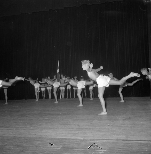 Schwarzes und weißes Foto einer Gruppe, die auf einer Bühne Gymnastik macht, mit einem Vorhang und einer Fahne im Hintergrund.