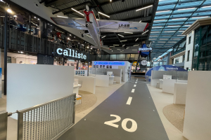 Ein großer Raum mit weißen Wänden, ein von der Decke hängendes Flugzeug, ein Geländer auf der linken Seite, Objekte auf dem Boden auf der rechten Seite, Texttafeln im Hintergrund und an Metallstangen an der Decke befestigte Lampen, wahrscheinlich am Düsseldorf Airport.