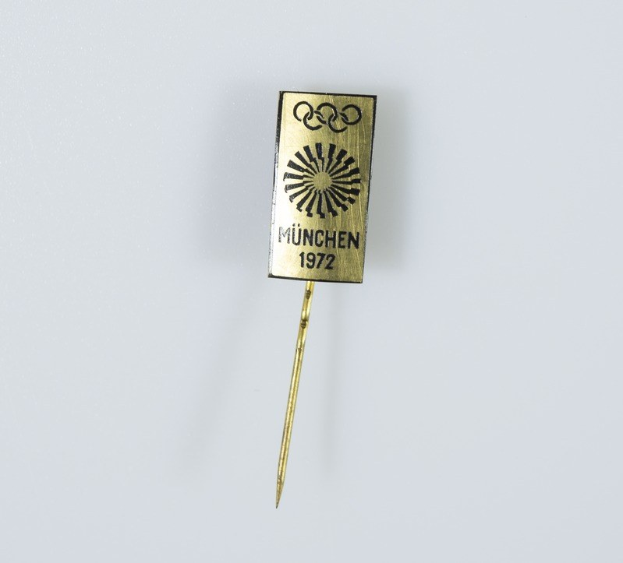 Goldene Nadel mit der Aufschrift "München 1972 Olympische Spiele" vor einem weißen Hintergrund.