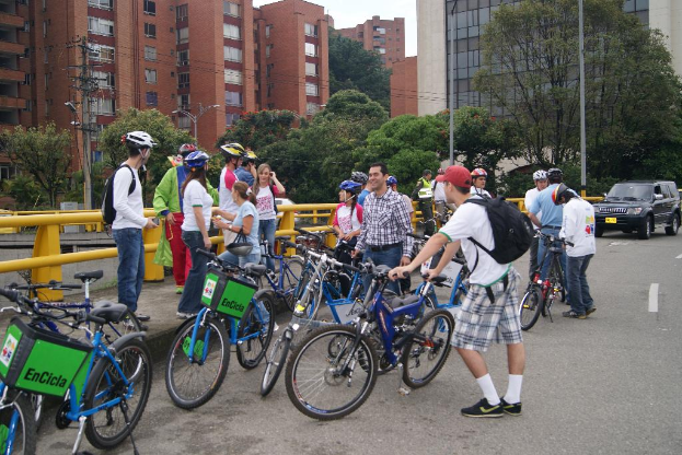 Eine Gruppe von Menschen, die meisten tragen Helme, fahren auf Fahrrädern auf einer Straße mit gelben Stangen, mit Pfosten, einem schwarzen Auto, Bäumen, Drähten, Gebäuden und dem Himmel im Hintergrund.