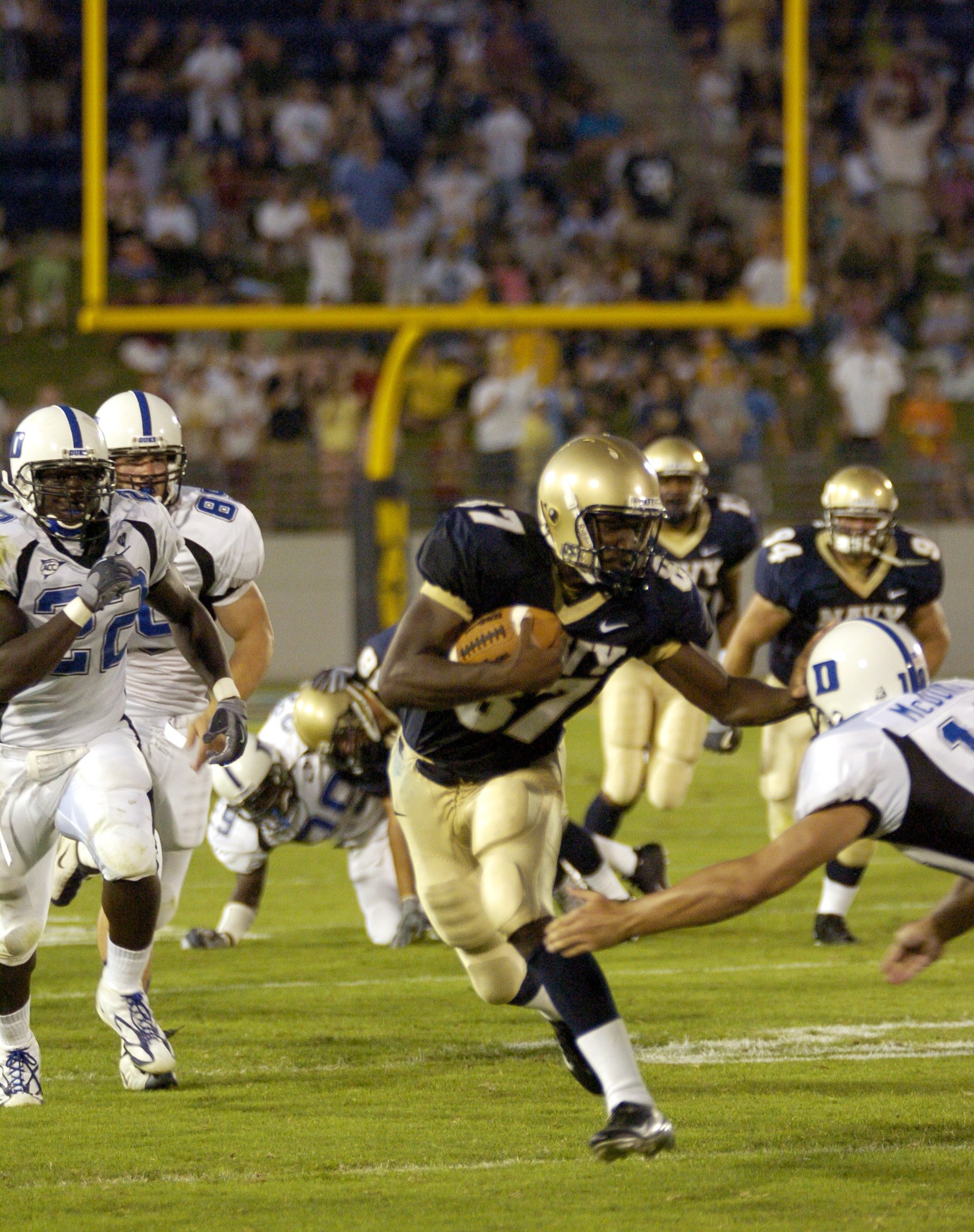 Fußballspieler in rotem Trikot mit Ball, verfolgt von Gegenspielern in Sportbekleidung und Helmen, mit unscharfen Zuschauern und Stadionelementen im Hintergrund.