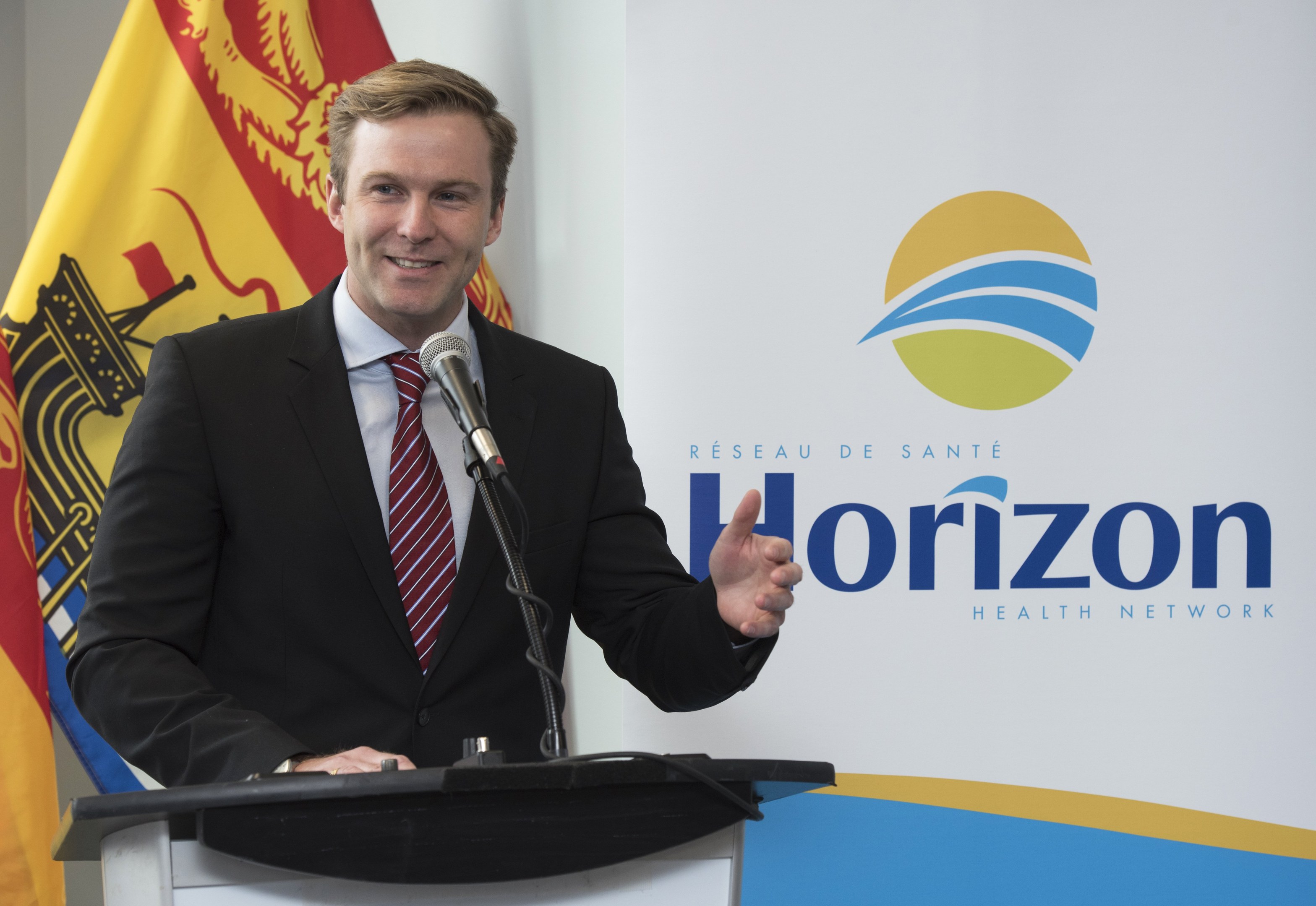 Ein Mann in einem schwarzen Blazer, weißem Hemd und roter Krawatte steht an einem Podium mit einem Mikrofon und einem Banner im Hintergrund und spricht als CEO.
