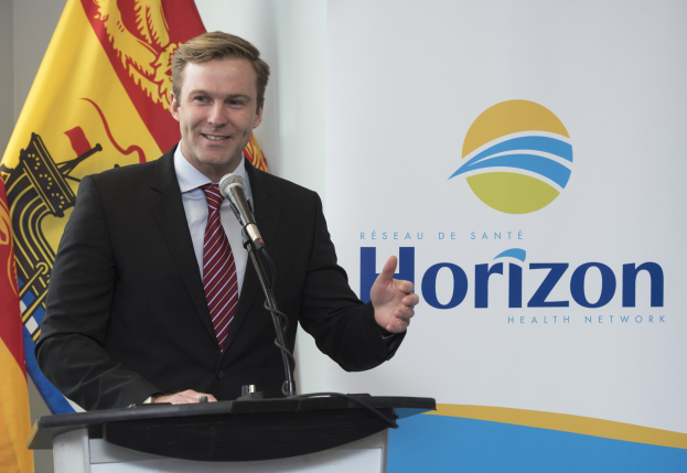 Ein Mann in einem schwarzen Blazer, weißem Hemd und roter Krawatte steht an einem Podium mit einem Mikrofon und einem Banner im Hintergrund und spricht als CEO.
