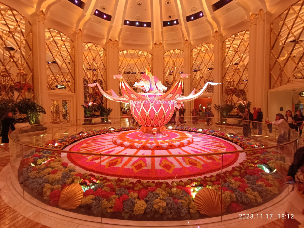 Außenansicht des Bellagio Hotels und Casinos in Las Vegas, Nevada, mit Menschen, dekorativen floralen Bodenmustern, beleuchteten Säulen und einem Wasserzeichen unten.