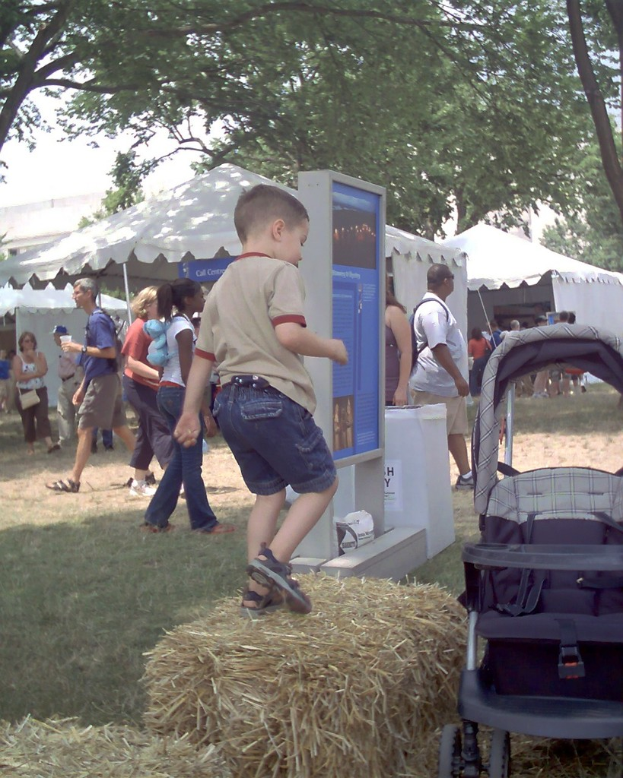 Eine Szene mit mehreren Zelten und Menschen, die herumlaufen, ein Displaybrett vorne, trockenes Gras mit einem Kind darauf und Bäume auf beiden Seiten.