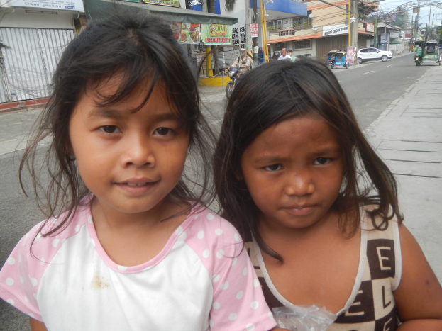 Zwei junge Mädchen stehen nebeneinander auf einer Straße mit Fahrzeugen, Menschen, Gebäuden, Pfählen, Drähten, Bäumen und Schildern im Hintergrund.