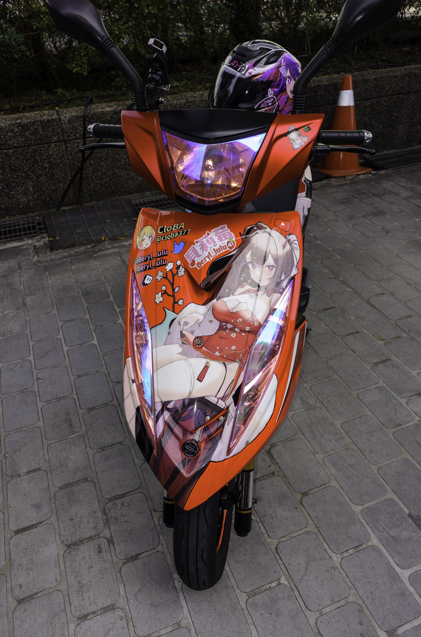 Ein oranger Roller mit Helm auf einer Stra√enseite neben einem Verkehrskegel, einer Wand und Bäumen.