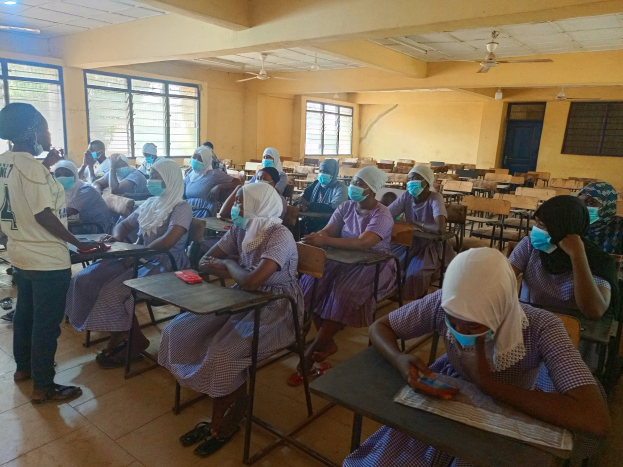 Ein Klassenzimmer in Nigeria mit Schülern, die Gesichtsmasken tragen und auf Bänken sitzen oder stehen, umgeben von Büchern und Gegenständen, mit Fenstern, einer Tür, Lampen und Ventilatoren im Hintergrund.