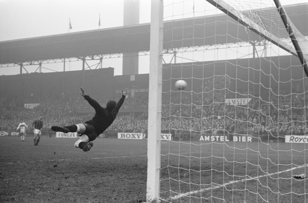 Ein Schwarz-Weiß-Foto eines Fußballspielers, der vor einem Tor einen Ball hält, mit einem Netz auf der rechten Seite und einer Zuschauermenge im Hintergrund, sowie Schildern mit Text, einer Brücke und Fahnen mit Stangen.