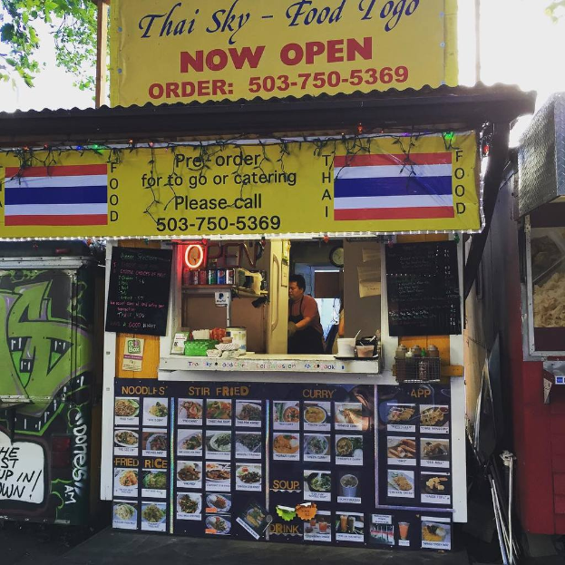 Ein Schaufenster mit verschiedenen Gegenständen, darunter Getränkedosen, Speisekarten, eine Uhr, Gewürze in Plastikbehältern, Gewürze, Werbetafeln, Namensschilder und ein Blick auf den Himmel.